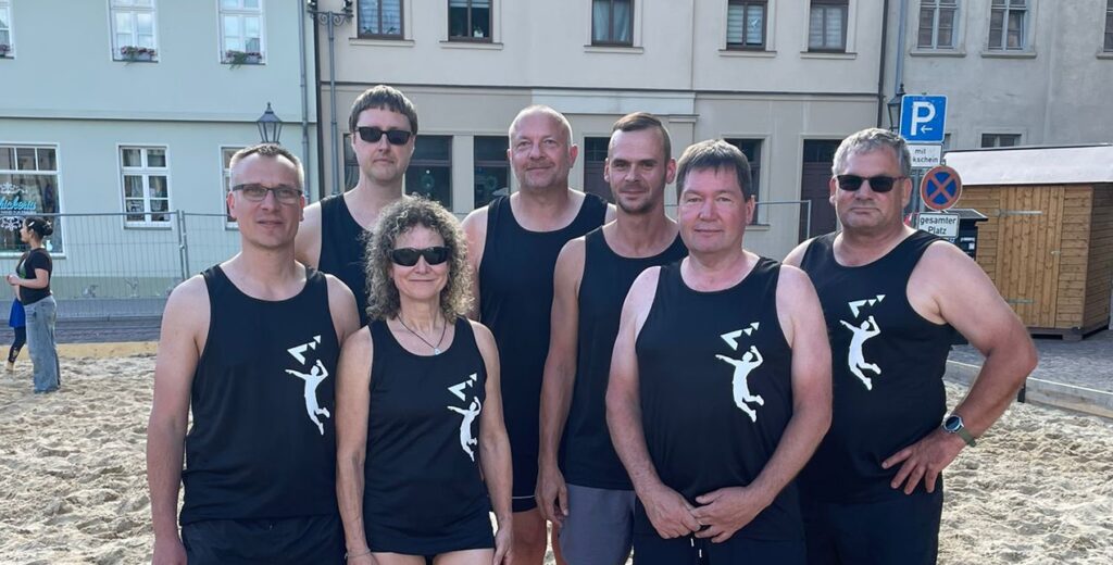 Sieben Personen in schwarzen Beachvolleyball-Trikots stehen auf Sandplatz vor Häuserfassaden.